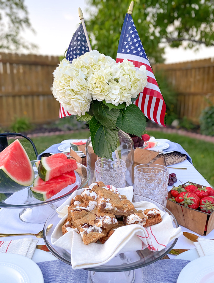 Red, white, and blue patriotic table set for a backyard BBQ party for Memorial Day with summer foods including smores bars, strawberries, watermelon, and mini brownie bites