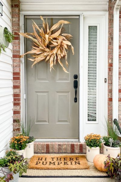 Small front porch decorated for fall with mums, pumpkins and a cornhusk wreath on the front door