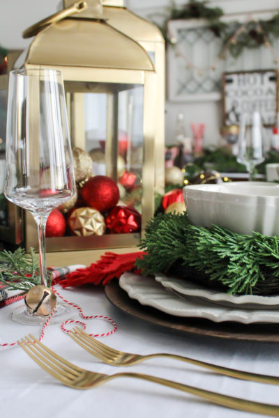 A Christmas place setting with red and gold decor, gold lanterns filled with Christmas ornaments, and a gold bell and greenry tied on the stem of a champagne glass