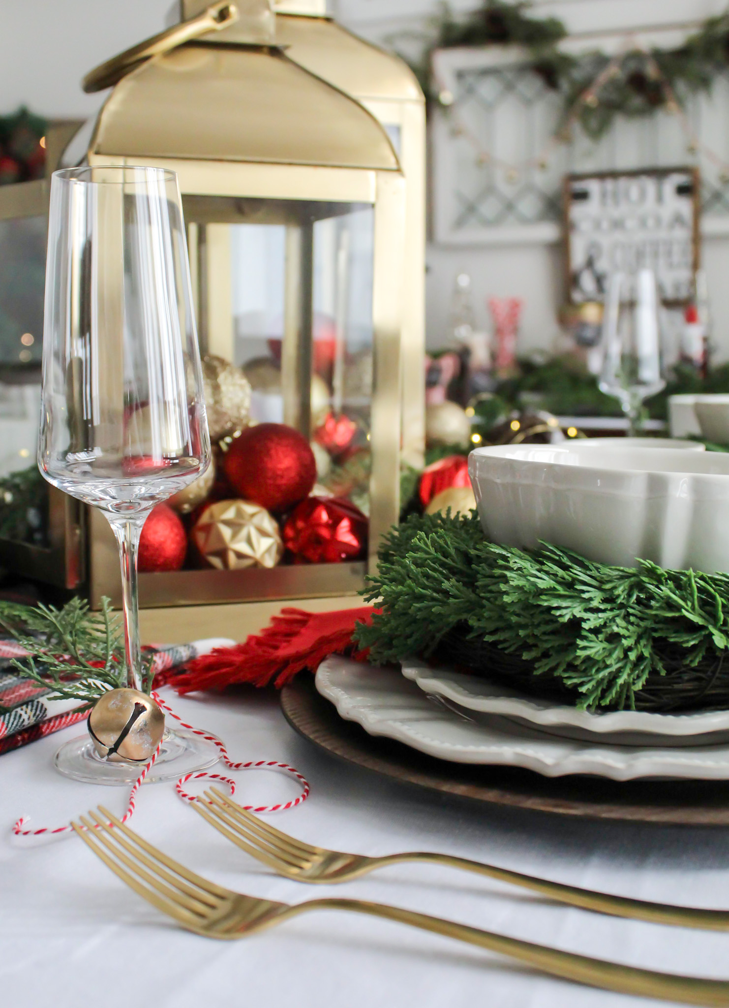 A Christmas place setting with red and gold decor, gold lanterns filled with Christmas ornaments, and a gold bell and greenry tied on the stem of a champagne glass