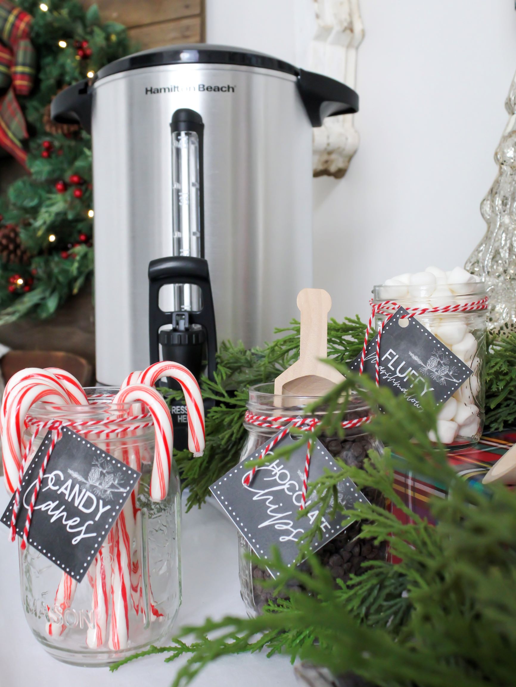 Large hot water dispenser on a holiday cocoa bar with hot cocoa toppings in mini mason jars