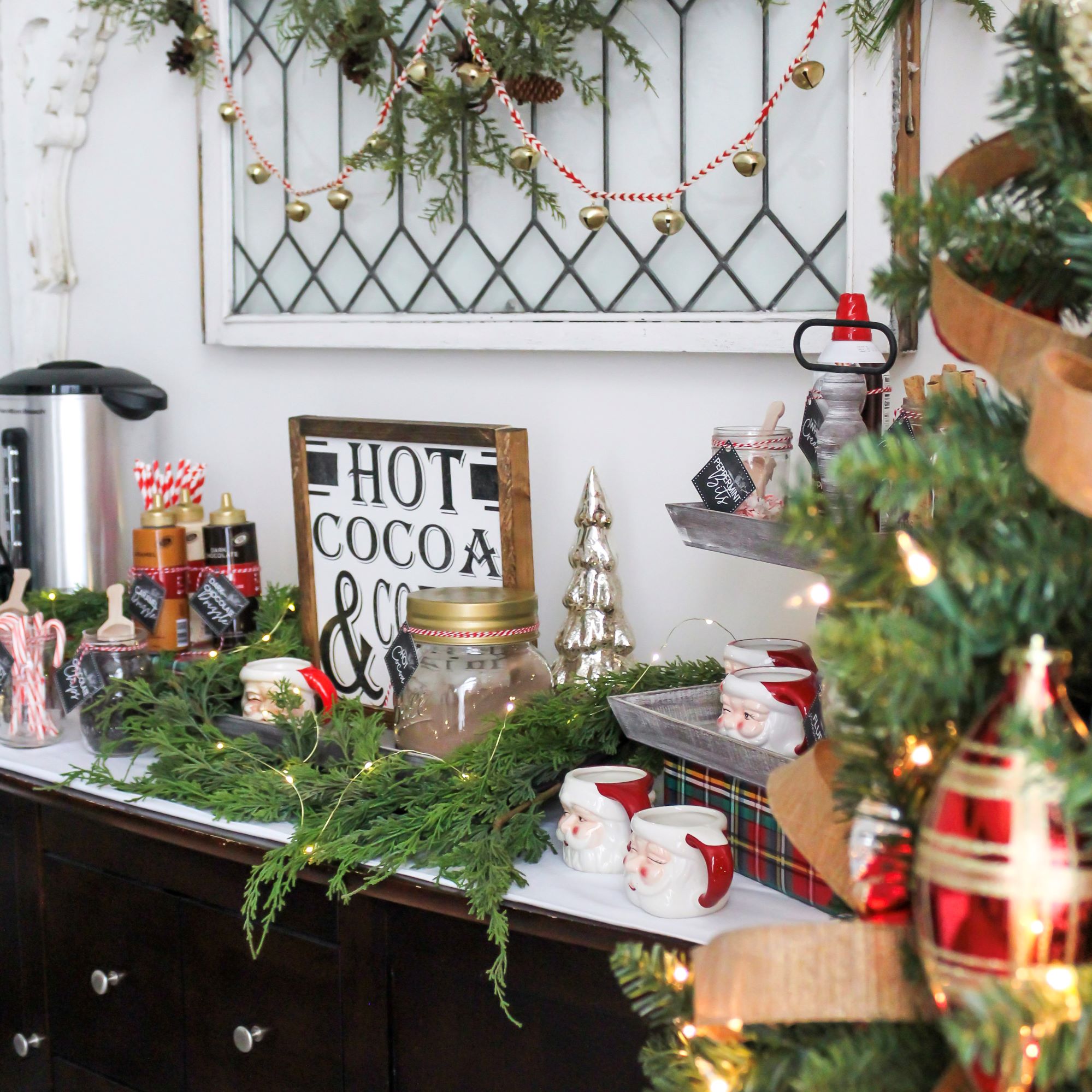 Cozy hot cocoa bar with all the toppings and with free printable tags set up on a buffet with Christmas decorations and a vintage window on the wall with holiday decor hanging above the buffet