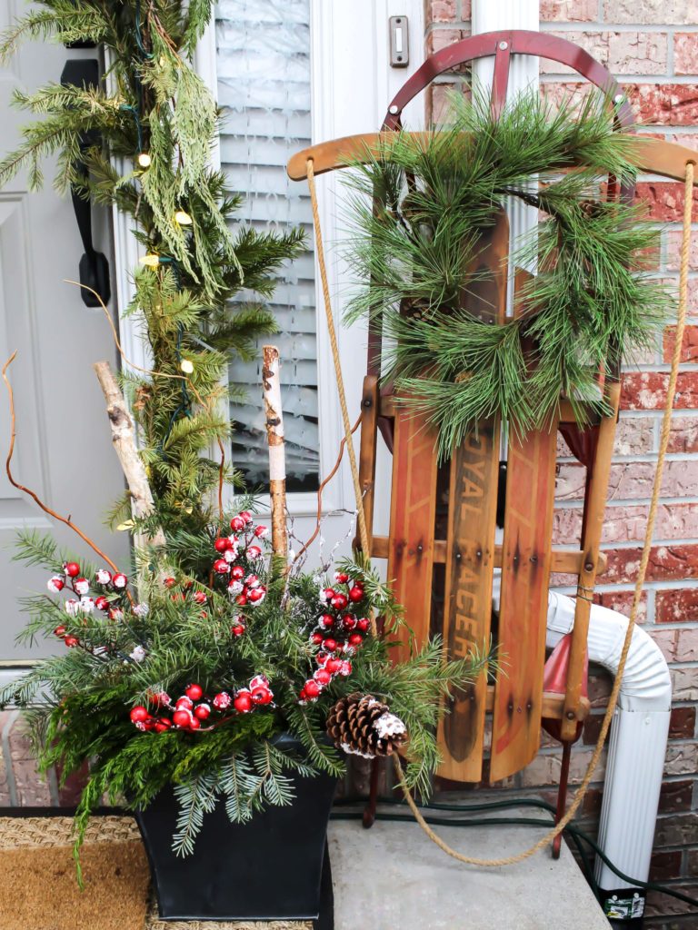 Front porch decorated for Christmas with a vintage sled leaned near the door and holiday planters filled with greenery, birch logs, and red holiday berries. 