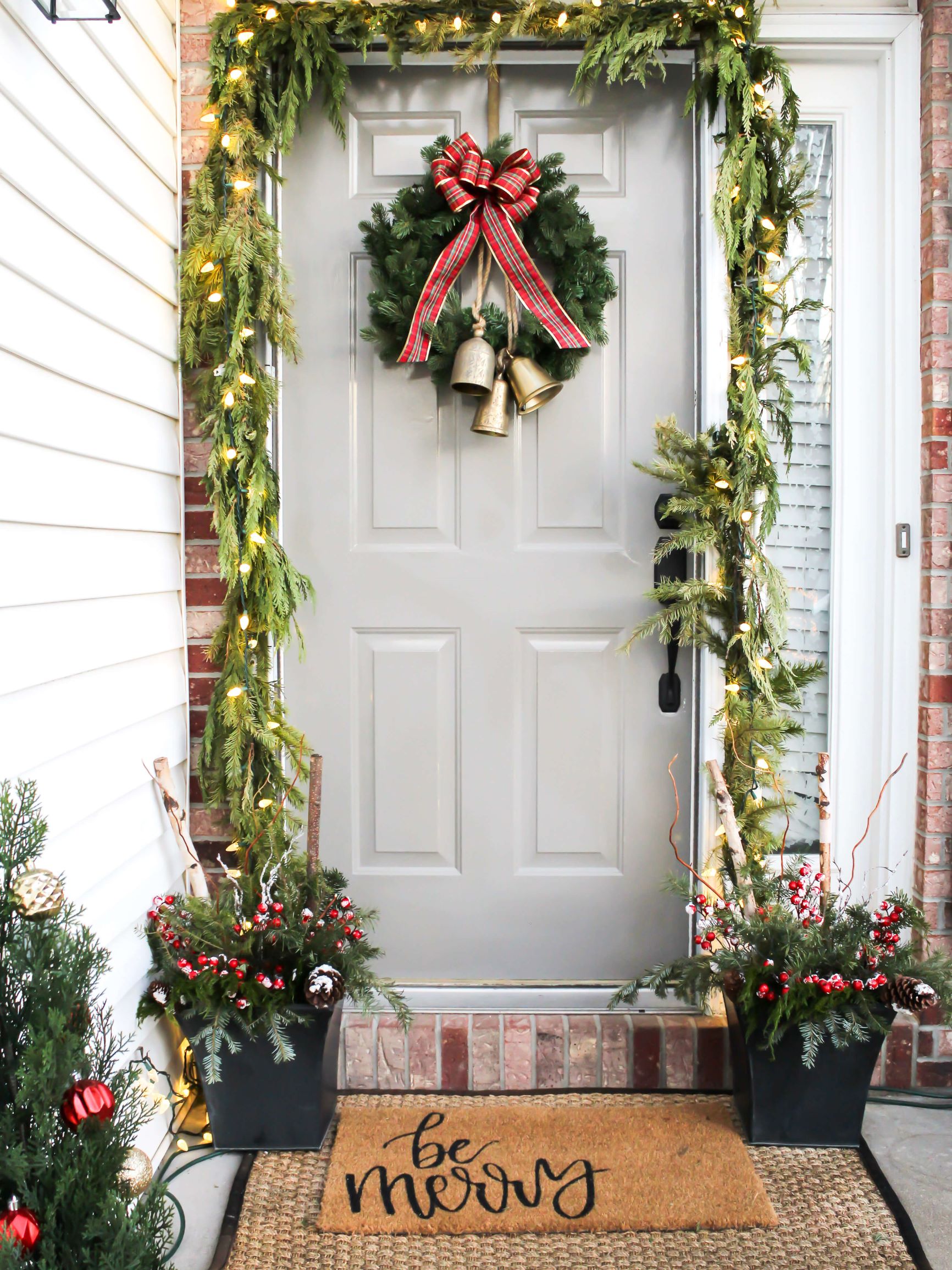 Front porch decorated for Christmas with garland draped around the door, a wreath with a bow and bells on the door, and holiday planters filled with greenery, birch logs, and red holiday berries. 