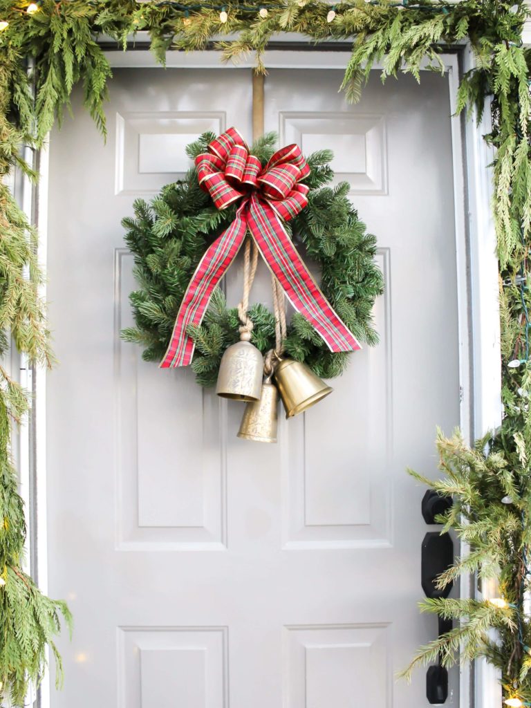 Front door decorated for Christmas with real garland draped around the door frame and a festive Christmas wreath with a red plaid bow and gold bells