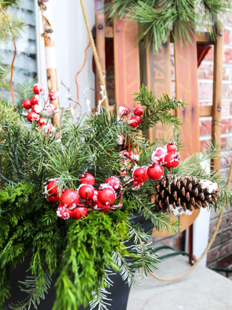 Up close view of holiday planters on the front porch filled with fresh greenery, pinecones, birch logs and red holiday berries. 