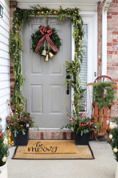 Decorating a small porch for Christmas with gold bells, garland, a wreath with a plaid bow, a vintage sled, and winter planters with greenery and red berries.