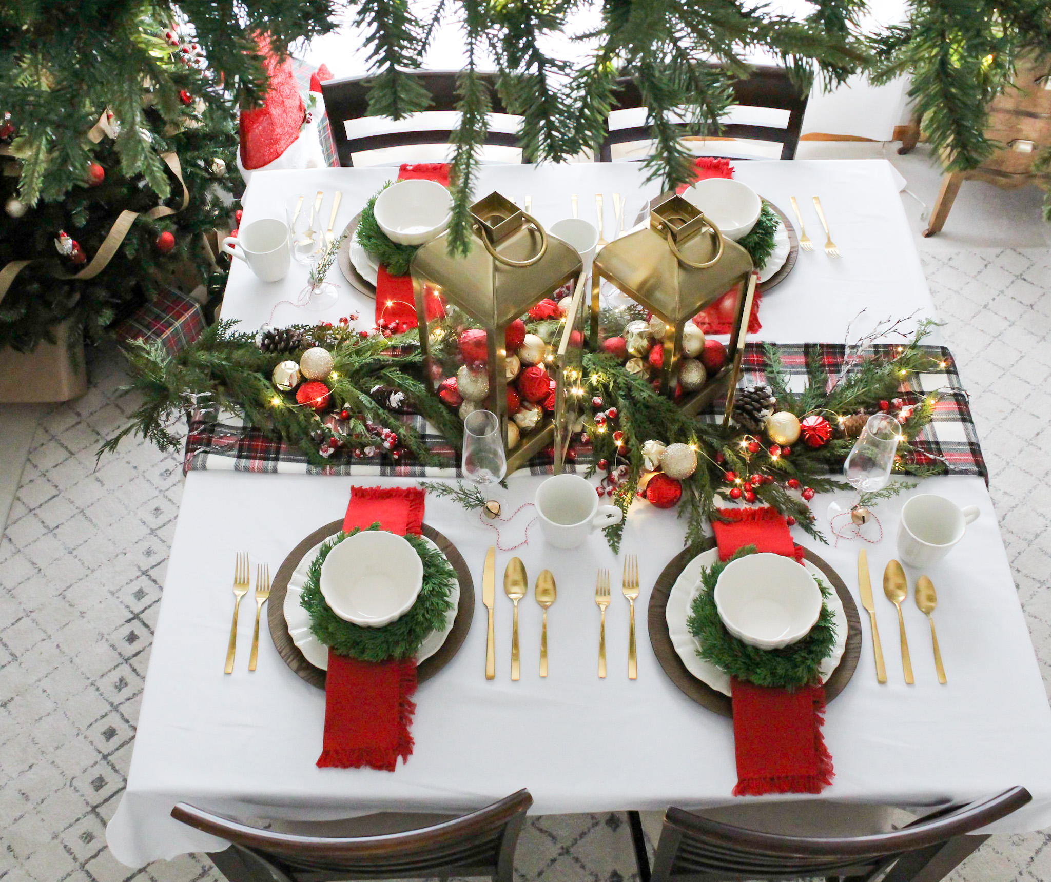 Overhead view of a dining room table set for Christmas brunch with red, white and plaid decor, Christmas greenery, twinkle lights, and large gold lantern cetnerpieces filled with ornaments.