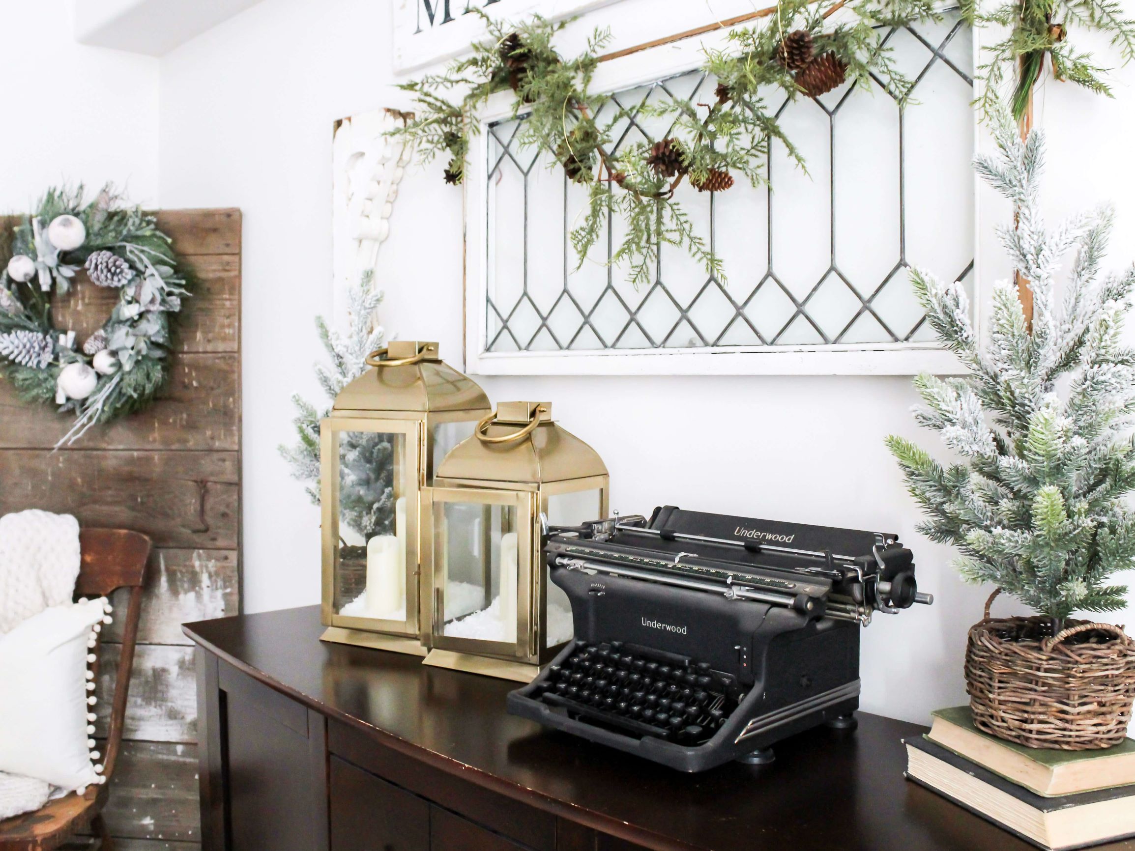 A neutral winter vignette styled on a buffet table in the dining room with gold lanterns and mini flocked trees in a basket with a barn doorn in the corner and a leaded glass window hanging on the wall above the buffet