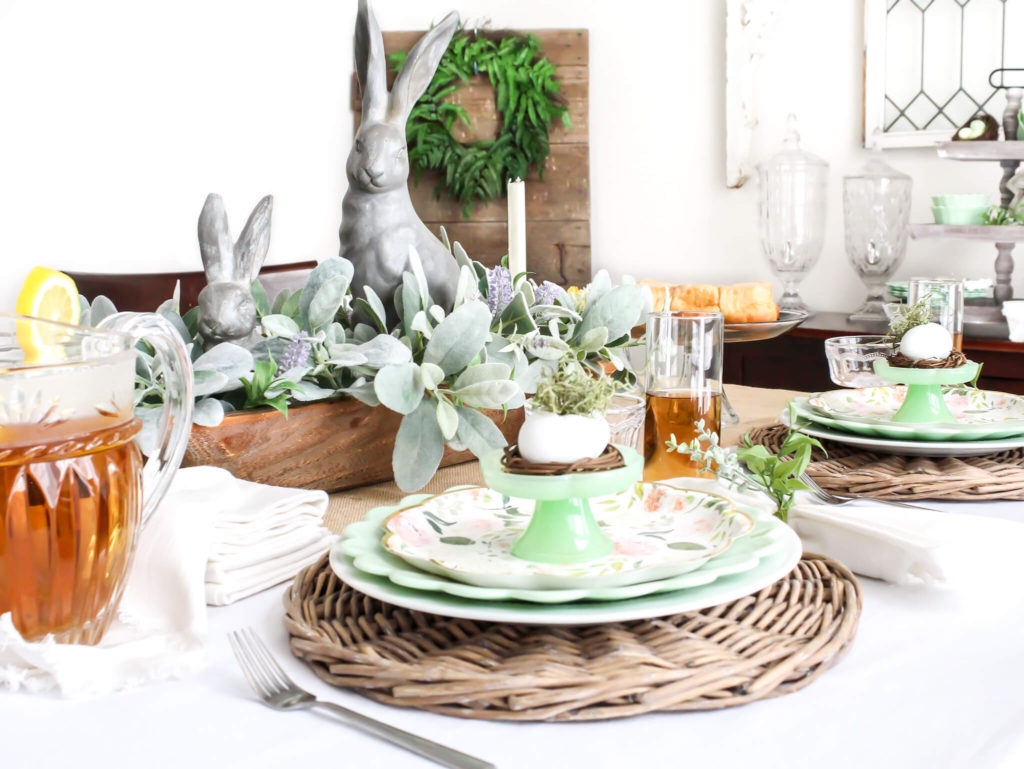 a dining room table set for Easter with jadeite dishes, floral paper plates, plastic easter eggs filled with moss and a springtime dough bowl centerpiece with bunnies and lambs ear