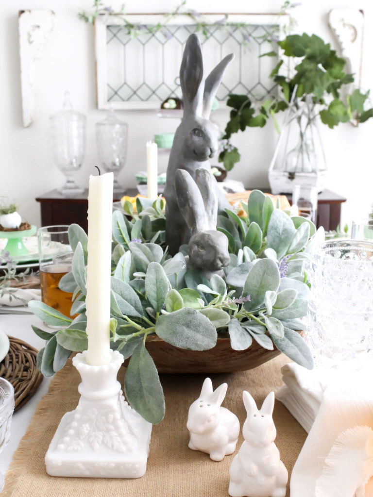 Close-up view of a spring centerpiece with a reproduction dough bowl styled with Pottery Barn Essex Easter bunnies, lambs ear greenery, and lavender and anqitue milk glass candle stick holders