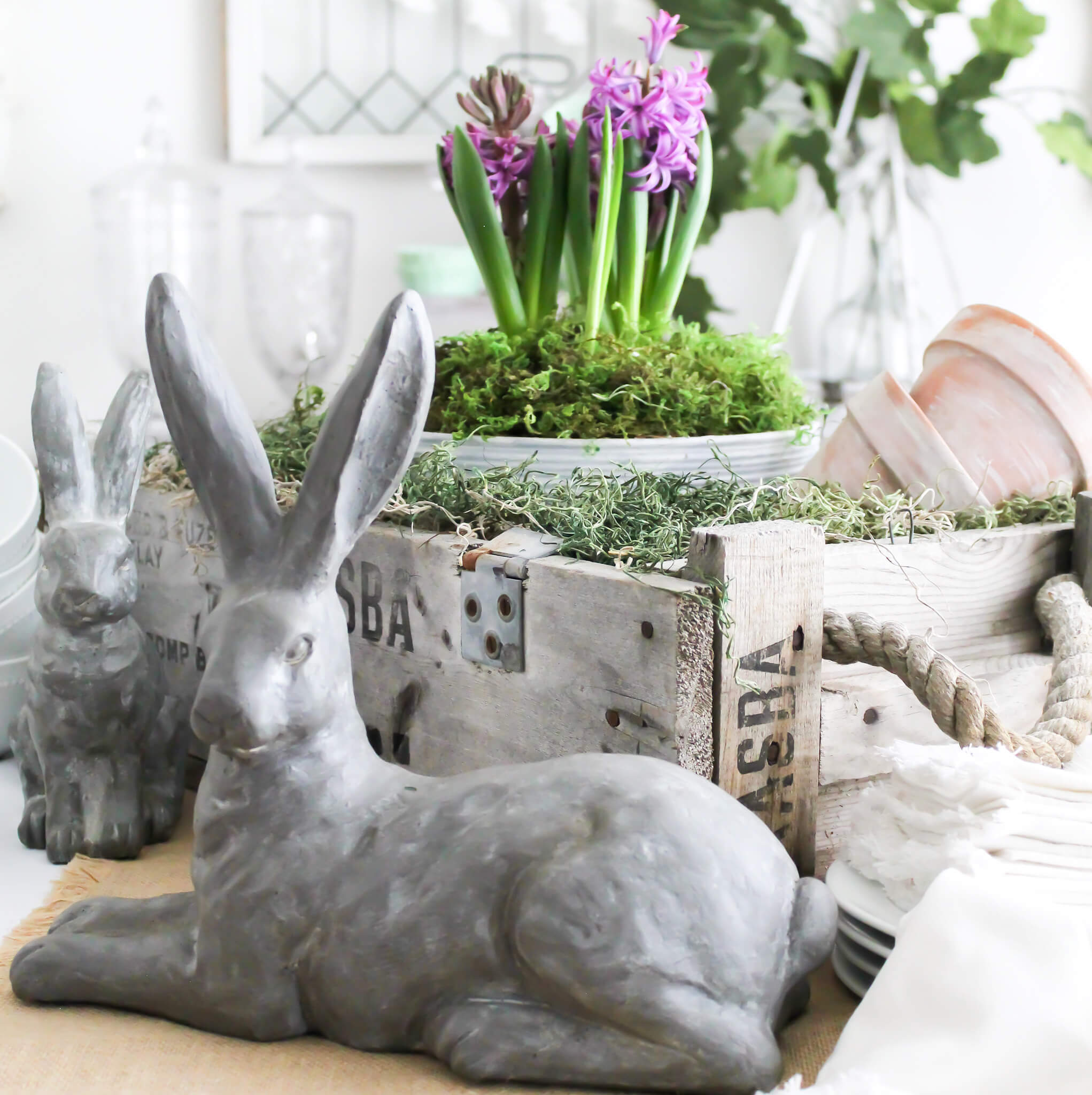 Aged terra cotta pots sitting in an old wooden crate on a dining room table during Eastesr. The larger pot is filled with moss and a purple hyacinth. Stone bunnies are in the foreground sitting on the table.