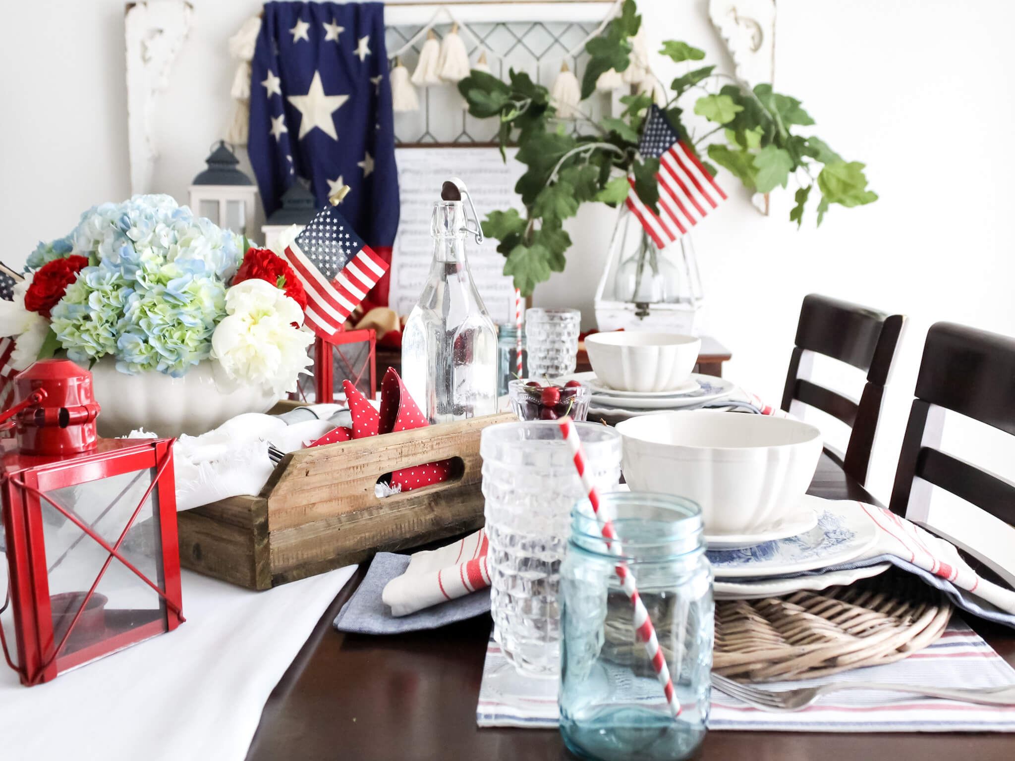 Patriotic 4th of July tablescape styled on the dining room table with red lanterns, american flags, and a rustic tray filled with a bouquet of red white, and blue flowers