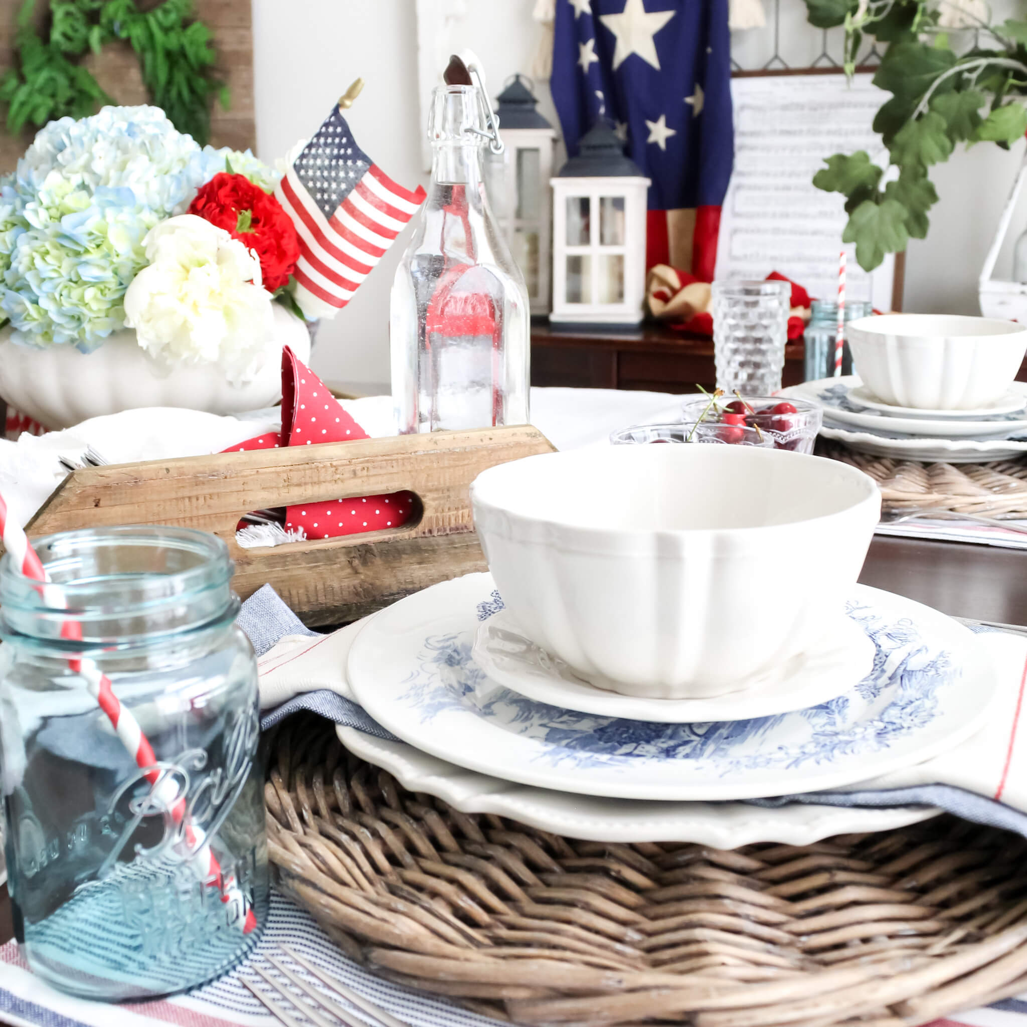 Fourth of July tablescape set on a dining room table with patriotic decor in the background including a vintage Amerian flag draped over an antique window hanging on the wall