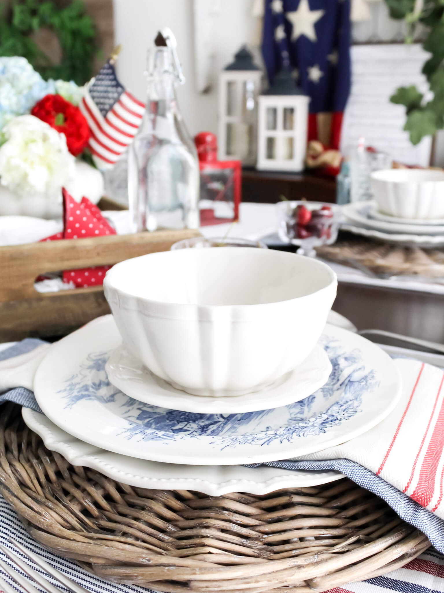 4th of July place setting created with patriotic decor including a round wicker charger, red and white striped napkins, blue transferware plates, and white dishes