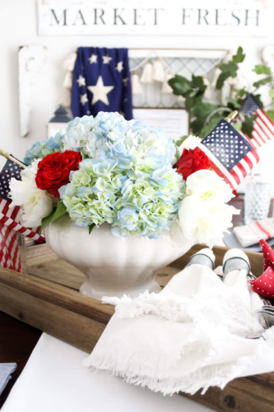 Patriotic 4th of July centerpiece with red, white, and blue fresh flowers and small American flags arranged in a vintage soup turee