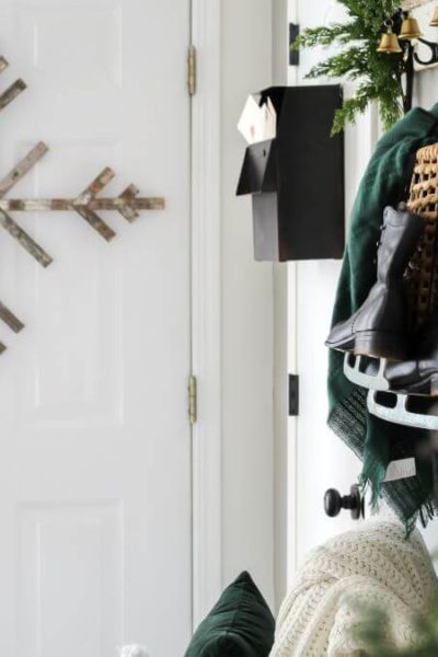 Entry decorated for Christmas with a wood snowfalk ornament hanging on the front door and a shelf with greenery and a baskt with a green blanket and old ice skates hanging from the hooks