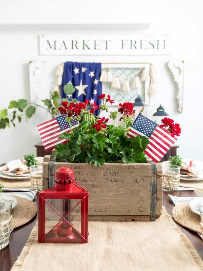 4th of July Centerpiece with Red Geraniums and American Flags in a Vintage Wood Blox Styled on a Dining Room Table with Vintage Window and Flag Draped on the Wall in the Background