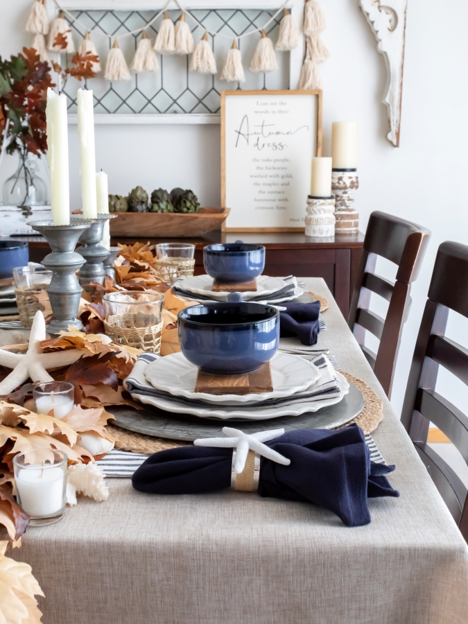 Long view of coastal fall tablescape with a leaf garland table runner, sea shell accents, blue and white dishes and rustic metal candlesticks