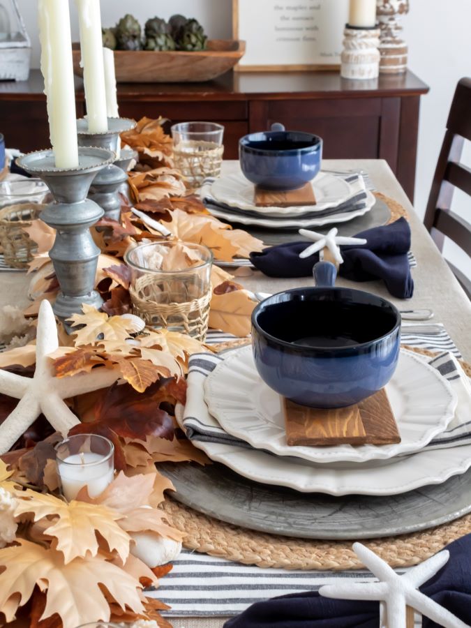 Close up view of coastal fall tablescape with a leaf garland table runner, sea shell accents, blue and white dishes and rustic metal candlesticks