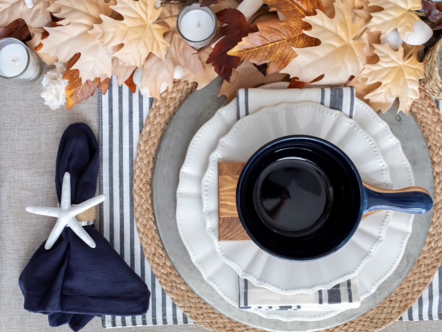 Overhead view of nautical and beach places setting for autumn with blue bowls, DIY coastal napkin ring holders, white plates, striped linens, and metal and woven chargers with a fall leaf table runner and sea shells. 