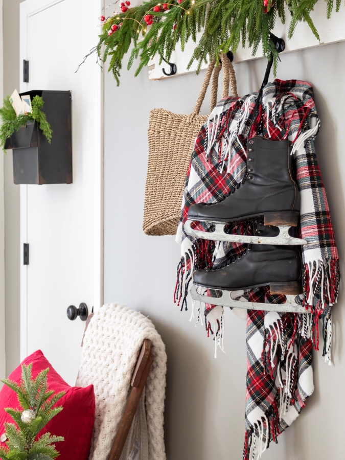 Entry decorated for Christmas wtih a maibox on a closet door filled with greenery and vintage postcards, and a shelf with a garland on it and a basket with a plaid blanket and vintage skates hanging from the shelf hooks