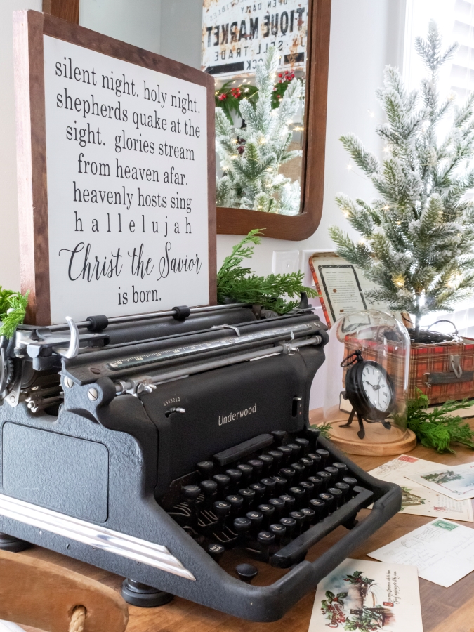 Christmas display set on a dresser incuding a vintage typewriter with a Christmas wooden sign, red plaid lunch box with a small tree placed inside and vintage postcards scattered on the dresser