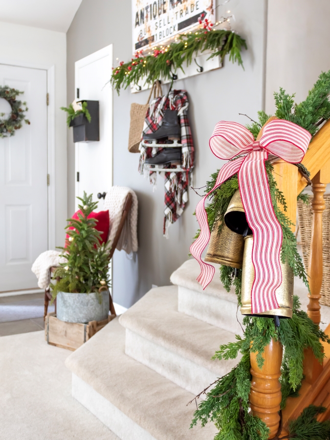 Entry decorated for Chrsitmas with red and white decor, a bow and bells on the stair railing, and vintage skates hanging from a shelf in the backgroud with a basket and plaid blanket