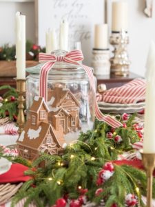 Glass jar with a ceramic gingerbread house inside, fake snow and bottle brush trees set in the center of a Christmas table setting with garland, twinkle lights, and frosted red berries