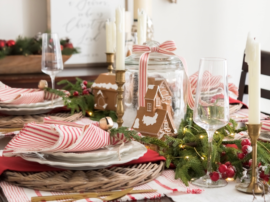 A Whimsical Red and White Gingerbread Holiday Tablescape with round wicker chargers, white dishes, red and white napkins, Christmas greenery and a gingerbread house centerpiece