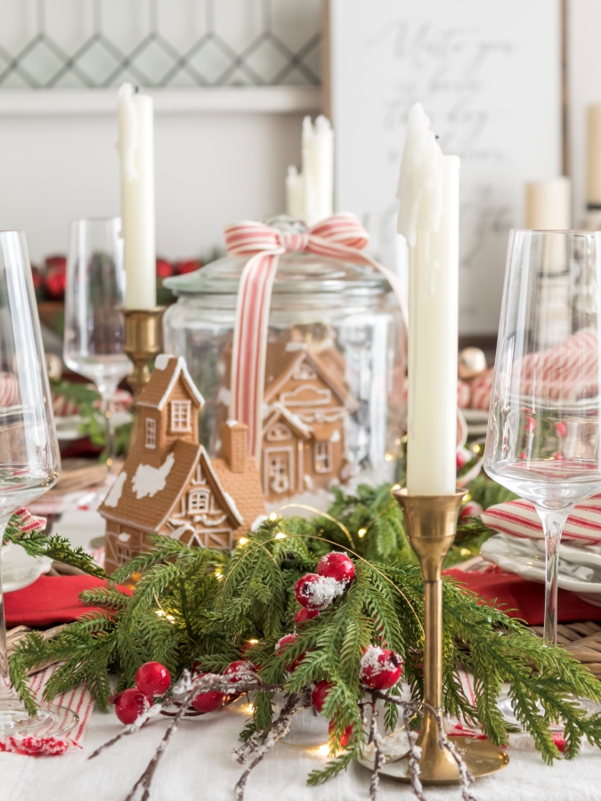 Red and white Christmas tablescape centerpiece with garland, red berries, ceramic gingerbread houses, and vintage brass candle sticks