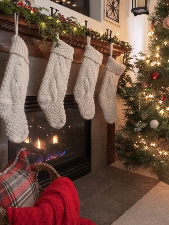 Christmas mantel with garland, white knit stockings, and a basket filled with plaid holiday pillows and extra winter blankets