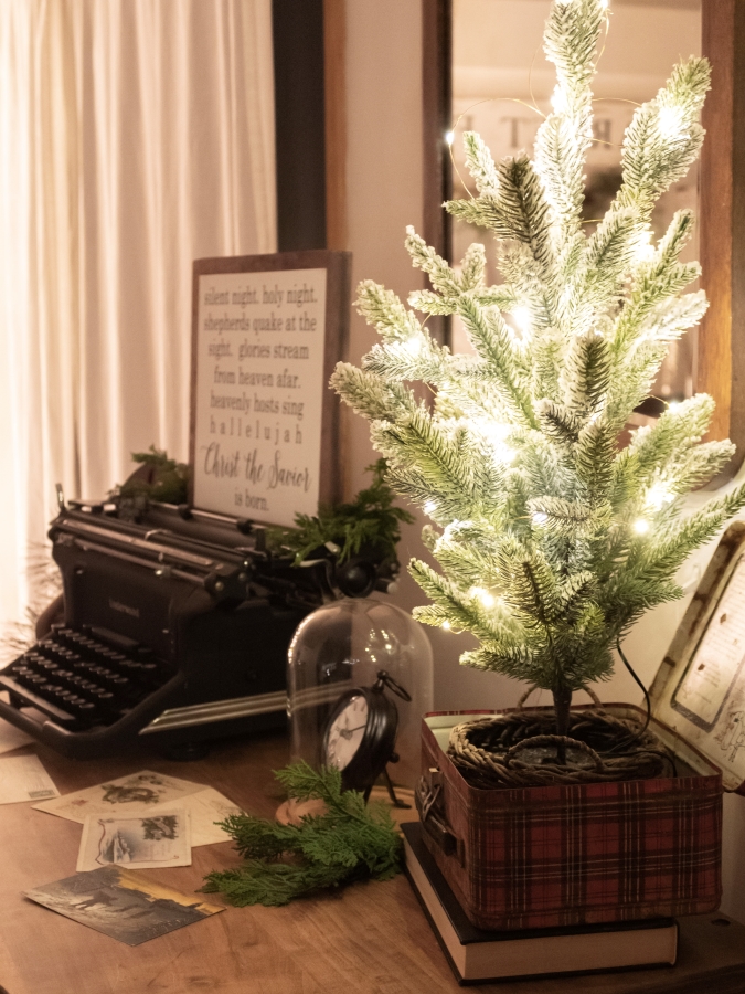 Small Christmas tree lit with twinkle lights displayed in a red plaid vintage lunch box on a dresser with a vintage typewrite and holiday sign in the background. 