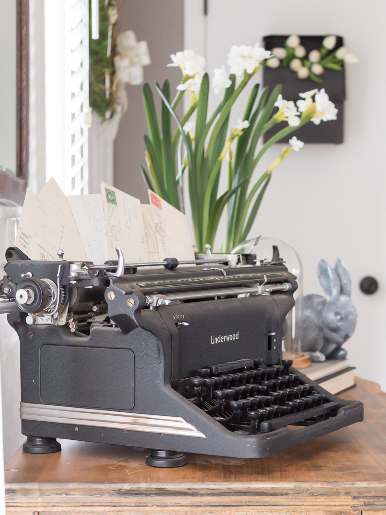 Spring vingnette with vintage typewriter and postcards, a bunny, and spring flowers in the background.