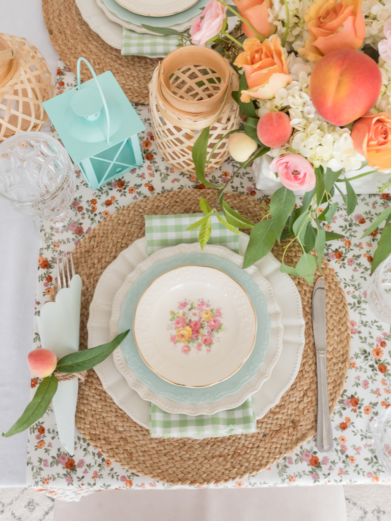 Overhead view of ummer cottage tablescape place setting with peach and mint green accents, vintage china, green gingham. napkins, a floral tablecloth, wicker lanterns, and fresh flowers with real peaches