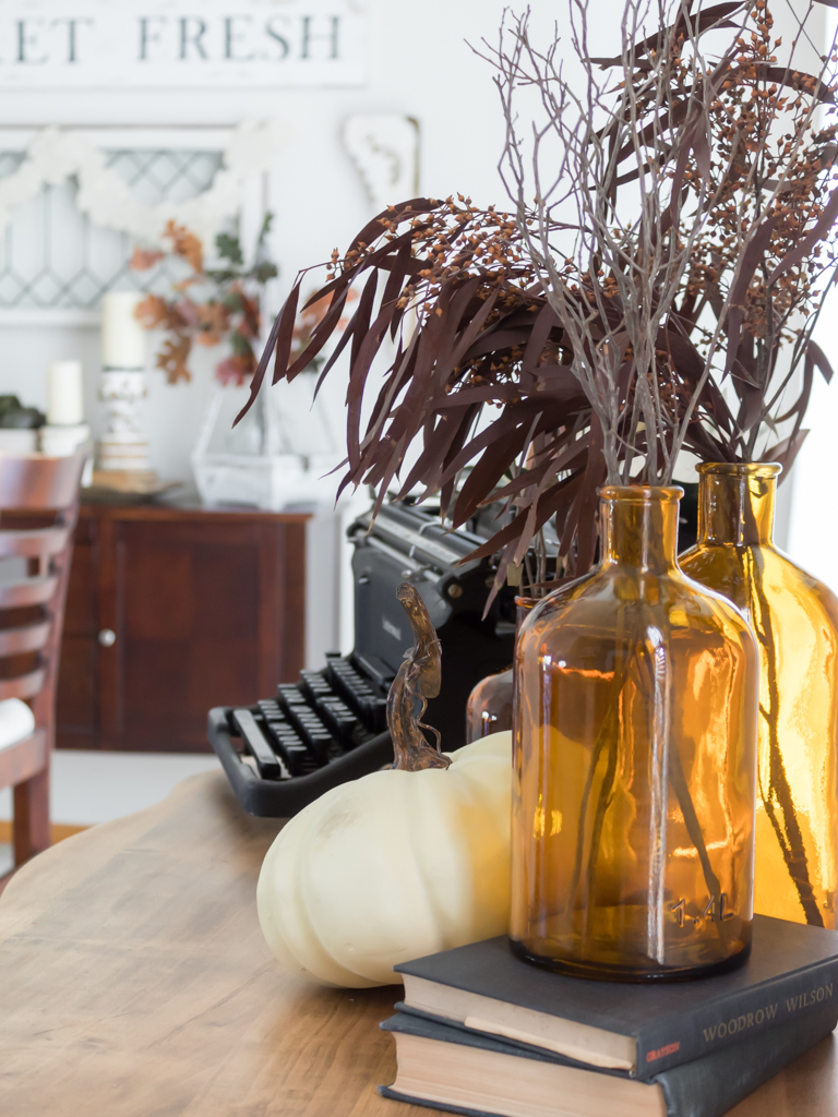 Amber bottles filled with fall foliage styled on a dresser with a vintage typewriter, pumpkin and old books