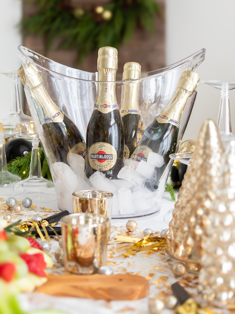 Mini bottles of champagne chilling on ice in a begerage tub set on a festive white and gold holiday table for a New Year's eve charcuterie party