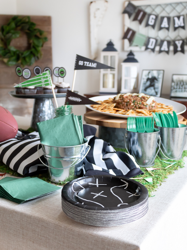 Black football party paper plates and green disposable napkins and silvereware in galvanaized buckets on a tablescape for a party for the Big Game