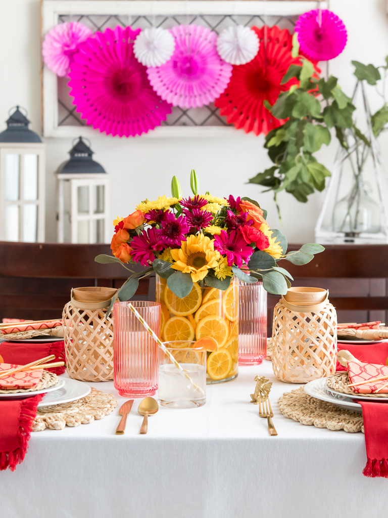 Dining Room Styled for a Chinese New Year Dinner Party with Bambo Lanterns, Woven Placemat with Red and Coral Linens and Brightly Colored Flowers with Sliced Oranges