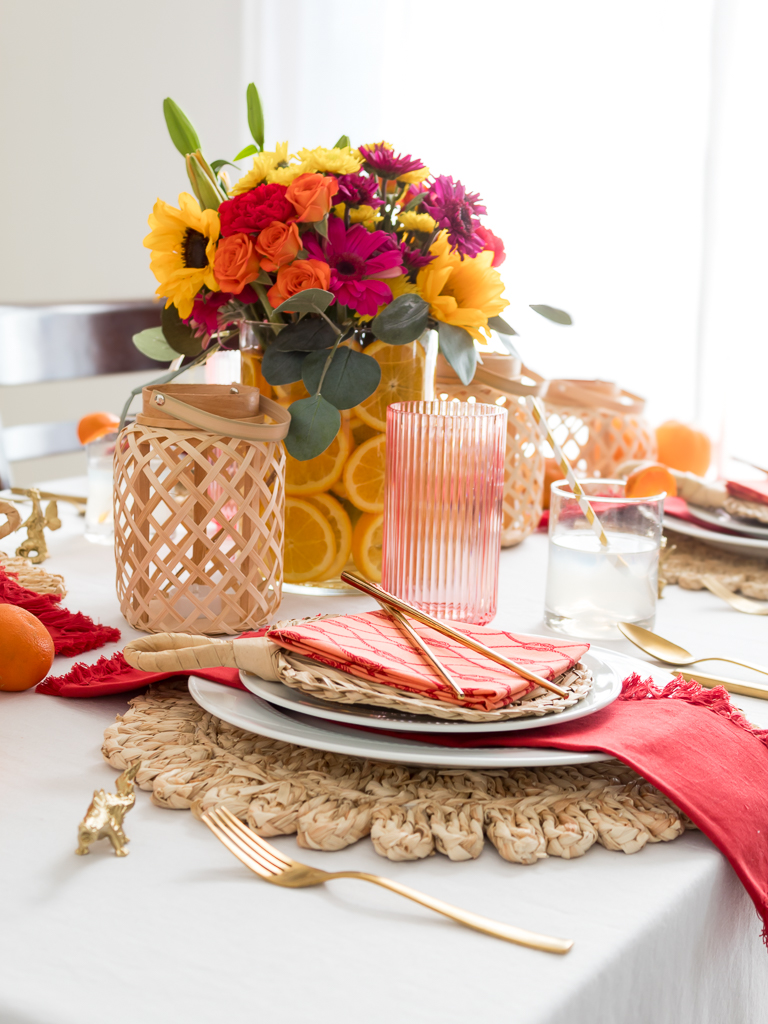 Chinese New Year Tablecape with Bambo Lanterns, Woven Placemat with Red and Coral Liensn and Brightly Colored Flowers with Sliced Oranges