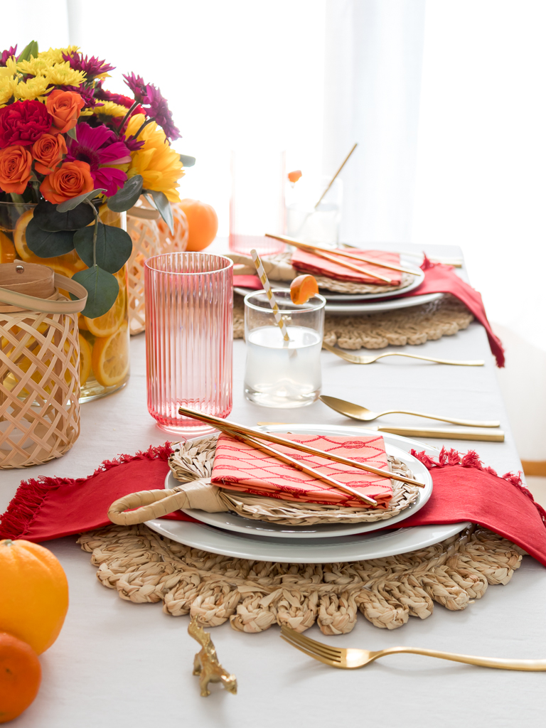 Tablescape Styled for a Chinese New Year Dinner Party with Bambo Lanterns, Woven Placemat with Red and Coral Linens and Brightly Colored Flowers with Sliced Oranges