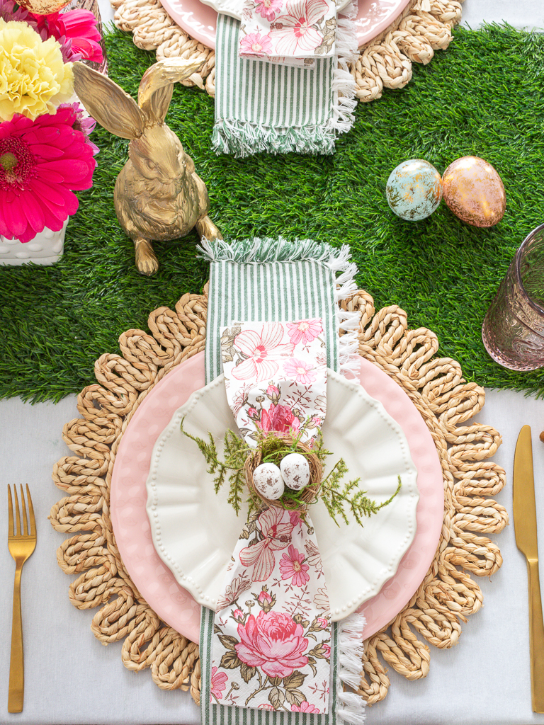 Overhead View of Pink, Green, and Gold Easter Place Setting with Round Woven Charger, Pink and White Plates, Floral Napkins, Green Ticking Stripe Napkins, Gold Silverware, and DIY Bird's Nest Easter Napkin Rings