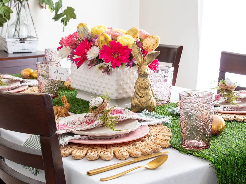 A close-up of a dining table set for an Easter dinner party. The table is decorated with colorful plates, folded napkins, birds nest napkin rings, pink glasses, gold siverware and a spring flower arrangement  in a hobnail vase with a bunny figurine in the center.