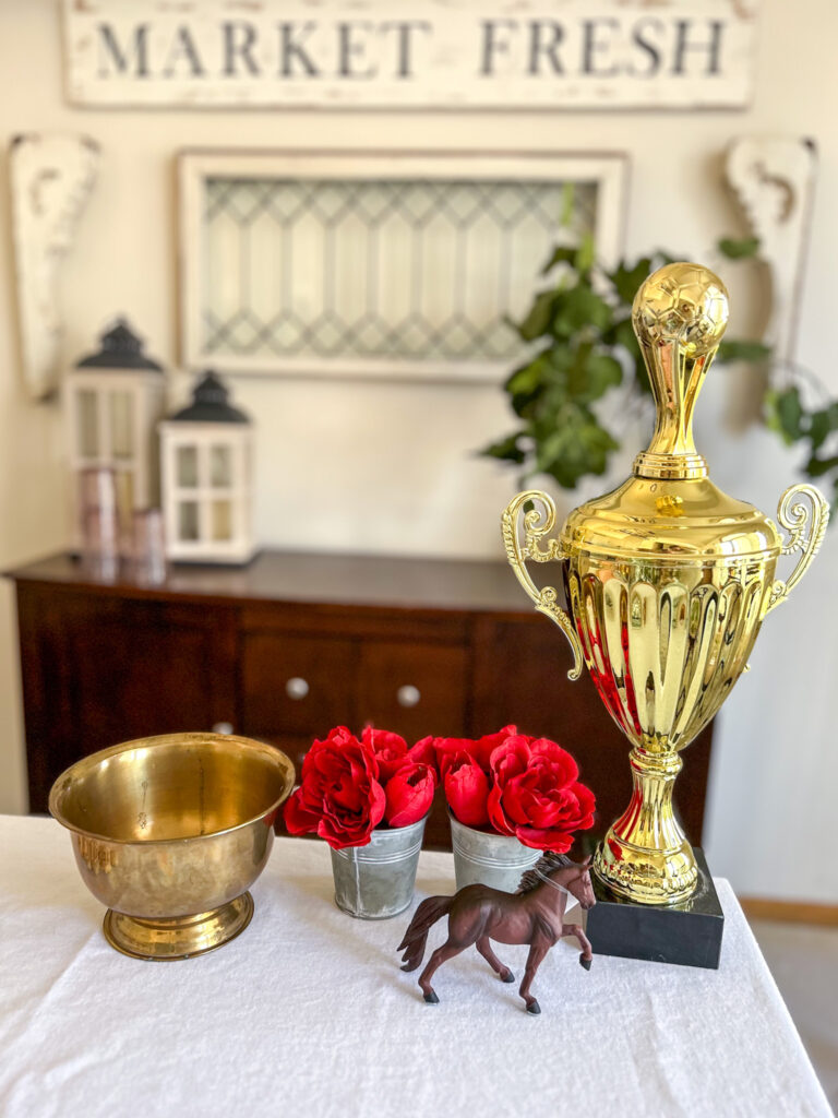 Thrifted decor items including, gold tropy, brass bowl, small tins with red silk flowers and toy horse for a Kentucky Derby DIY centerpiece