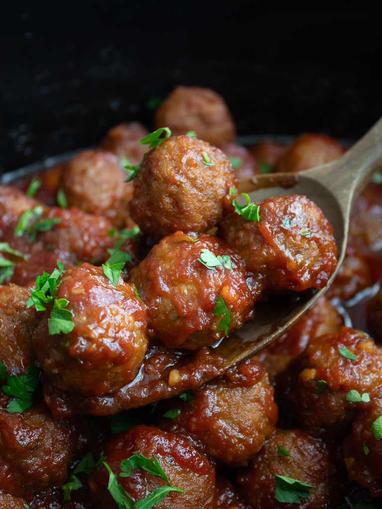 Close up view of Crockpot Bourbon BBQ Meatballs in the slow cooker displayed on a wooden spoon -Game Night Snacks