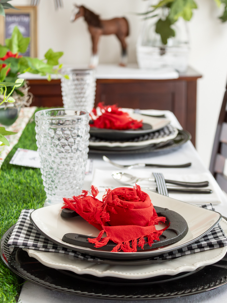 Close up view of a red and black place setting for a Kentucky Derby party with a black charger plate, gingham napkins, hobnail drinking glasses and a horsehoe and red napkin folded like a rose