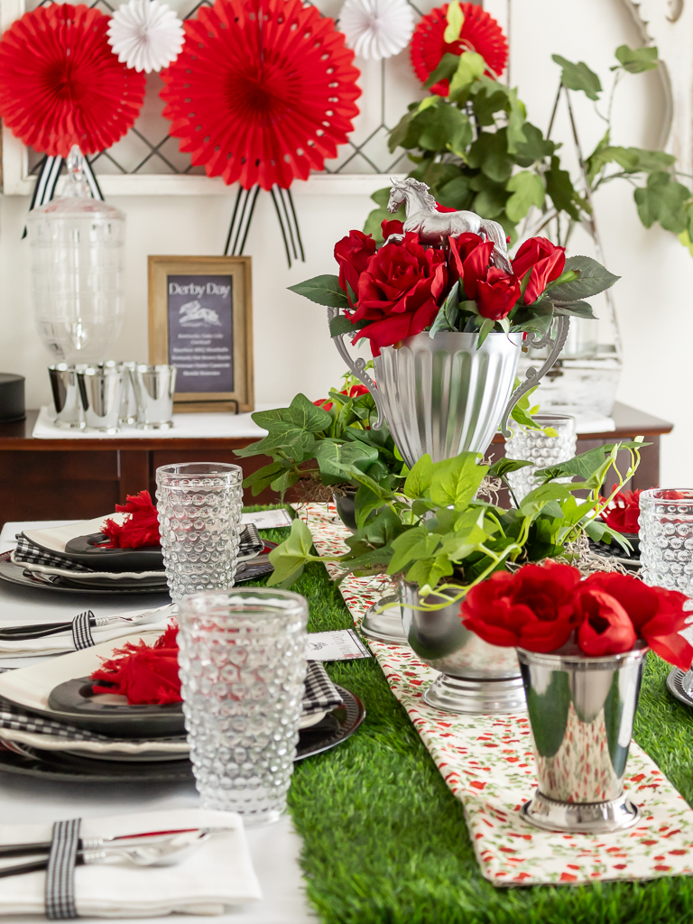 Centerpiece ideas for a Kentucky Derby party with a horse trophy filled with red roses, mint julep cups with red flowers and a green grass table runner