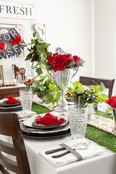 Side view of a dining room table styled for a Kentucky Derby-themed party with red and black decorations, a DIY trophy centerpiece with red roses, and mint julep cups filled with red flowers