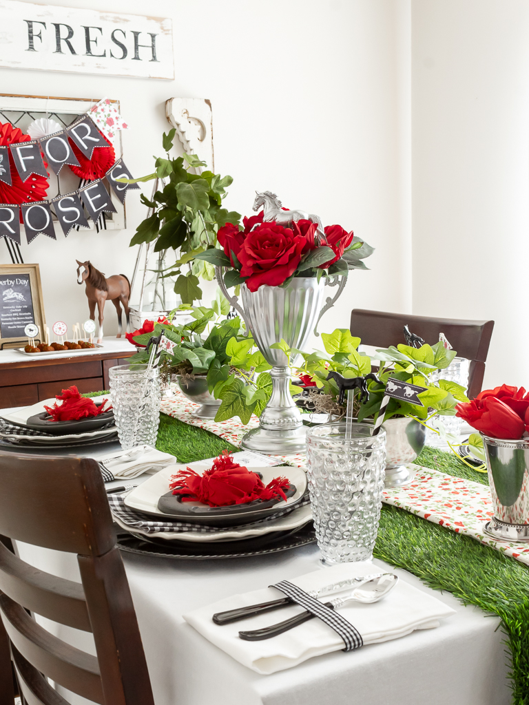 Side view of a dining room table styled for a Kentucky Derby-themed party with red and black decorations, a DIY trophy centerpiece with red roses, and mint julep cups filled with red flowers