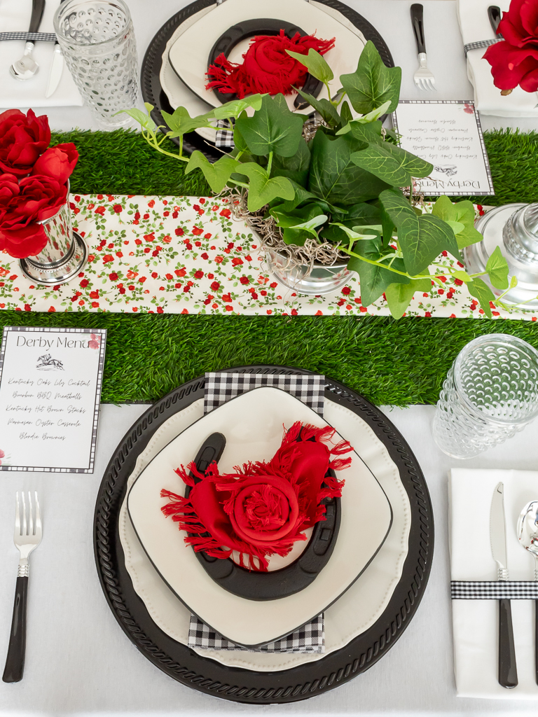 Overhead view of a red and black place setting for a Kentucky Derby party with a black charger plate, gingham napkins, hobnail drinking glasses and a horsehoe and red napkin folded like a rose